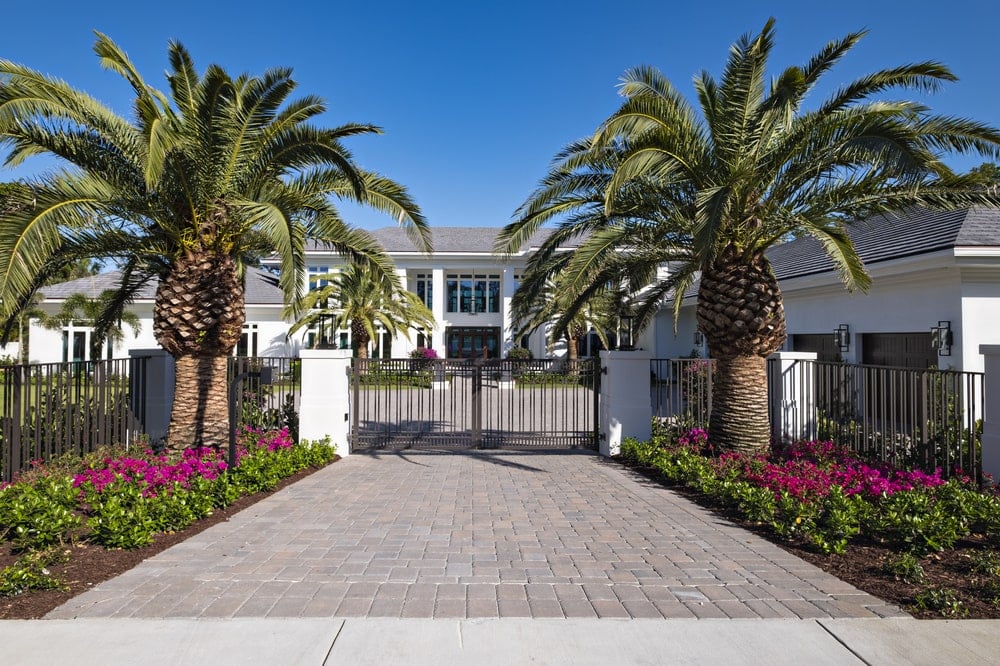This is a view of the house's main gate with a brick driveway flanked by colorful shrubs and large tropical trees.