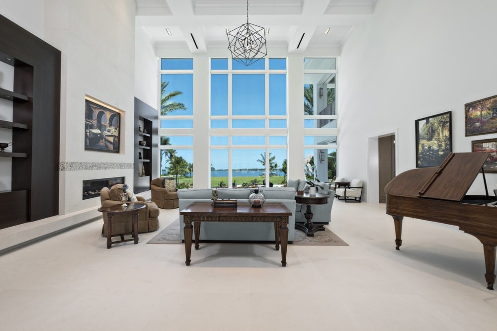 This is the large living room with a tall coffered ceiling, decorative chandelier and a modern fireplace with a wall-mounted artwork above across from the gray sofa set.