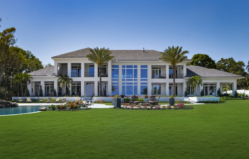 This is a view of the back of the house that has large glass walls and windows looking over a large grass lawn with tropical trees and shrubs.