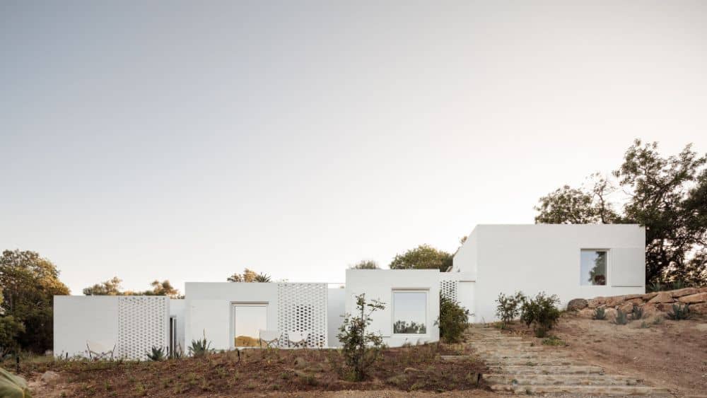 This is a full view of the adobe house with white exterior walls and large entryways complemented by the landscaping of shrubs and stone walkways.