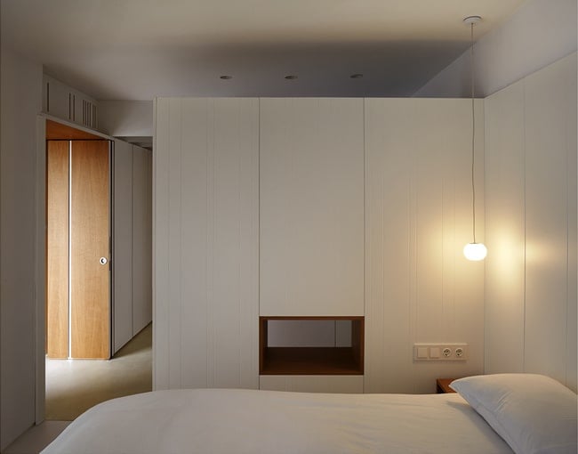 This is another look at the bedroom that has a white wall and a small window on the bottom part of the wall beside the bed along with a simple pendant light.