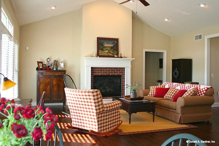 The living room has a vaulted ceiling, a brick fireplace, and a skirted sofa accentuated with red checkered pillows matching with the rocking chair.