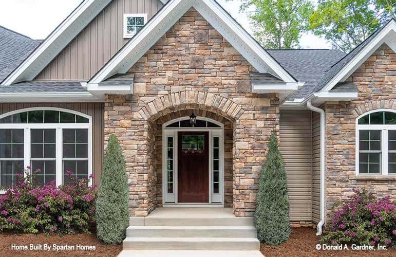This is a close look at the main entrance of the house with a mosaic stone brick exterior wall and archway flanked by medium-sized trees and colorful flowering shrubs by the sides of the walls.
