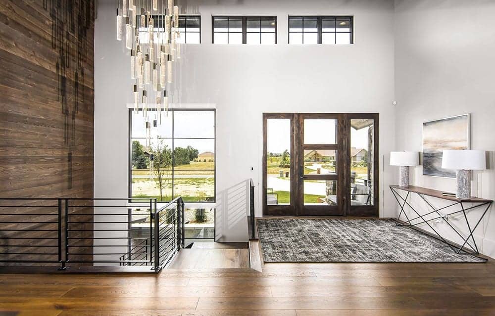 This is the mountain-style foyer with a tall ceiling, a row of transom windows, large windows and glass doors paired with a thin and tall console table on the side.