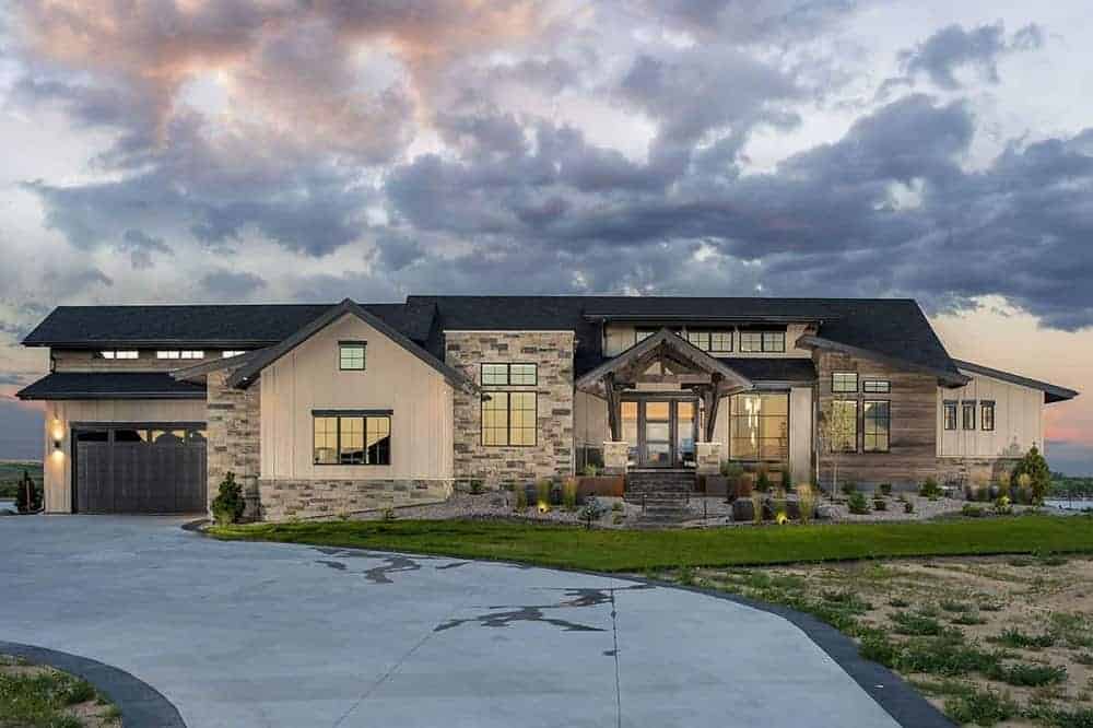 This is a look at the front exterior of a mountain-style home with a large concrete driveway, a grass lawn and pebbled planters on the side of the main entrance with shrubs and decorative rocks.