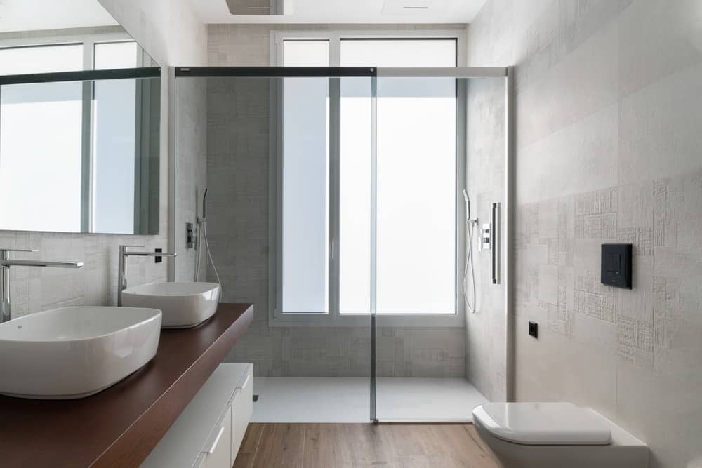 This primary bathroom has a floating two-sink vanity with a brown wooden counter that matches the floor. On the far side is the glass-enclosed shower area with a large window.