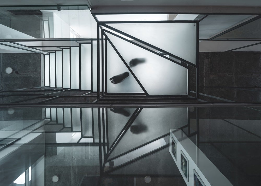 This is a look up the staircase tha has geometric wrout iron railings and frosted glass steps and has a glass wall on one side.