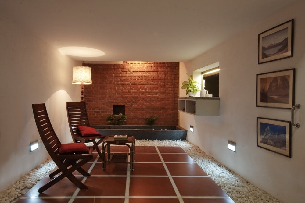 This is a comfortable room with a brown tiled flooring bordered with pebbles that complements the pair of wooden chairs. On the far side is a brick wall with a water feature.