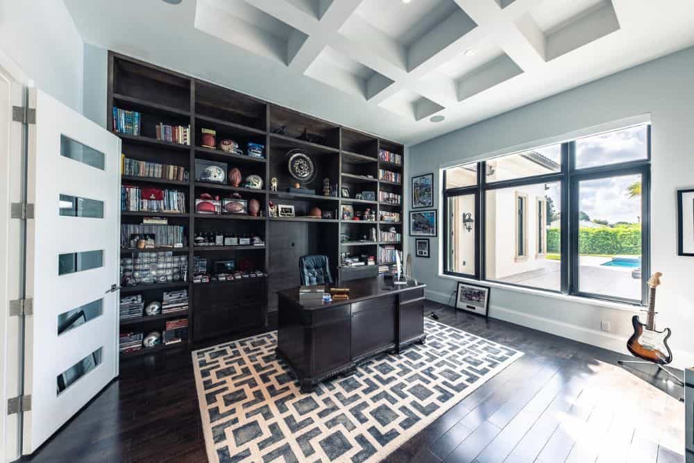 This is the large home office with a dark wooden desk that matches the dark wooden built-in shelves on the wall and the hardwood flooring topped with a coffered ceiling.