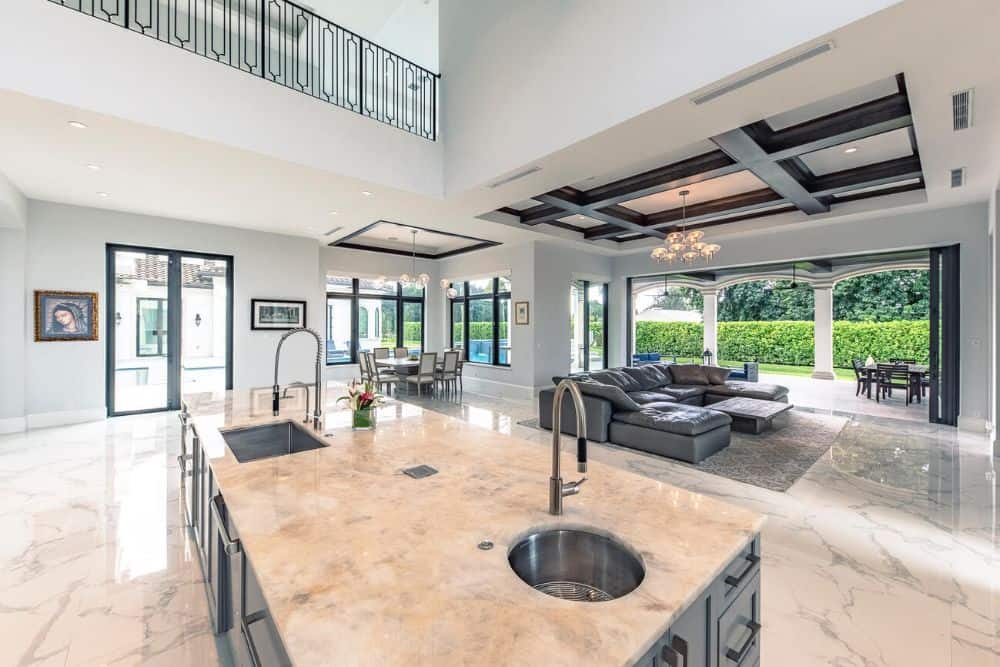 This is a close look at the kitchen island with a clear view of the family room with its large gray sectional sofa facing the TV topped with a coffered ceiling and a chandelier.