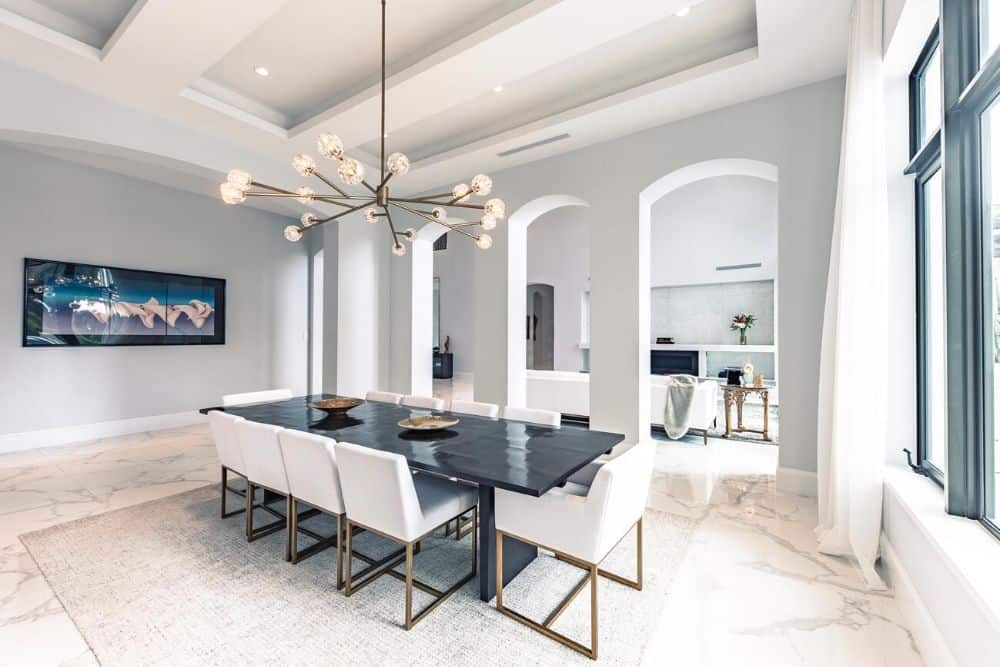 This is a close look at the dining room that has arched entryways, white coffered ceiling, a decorative chandelier and a dark dining table contrasted by the white cushioned chairs.