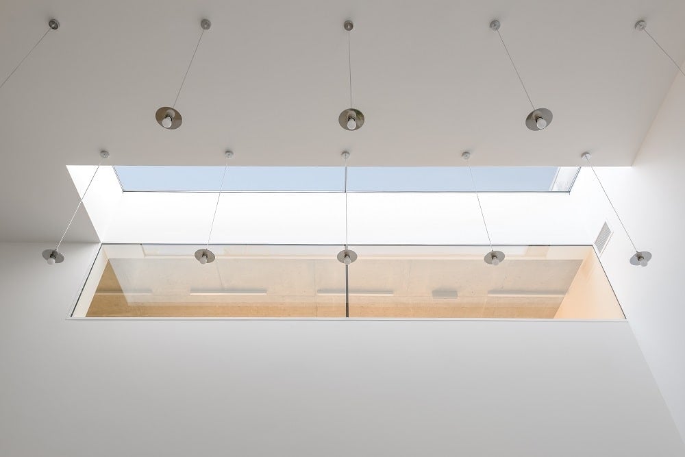 This is a look at the ceiling of the top hallway with a skylight and a glass window. There are also rows of pendant lights to brighten the white walls.