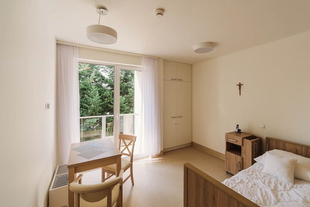 This is a look at the bedroom with a large wooden bed with a bedside table and a small table and chair area by the window.
