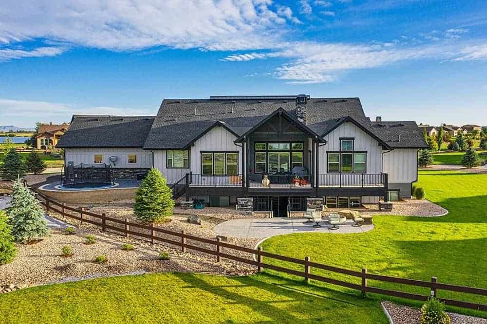 This is an aerial view of the back of the house that has glass walls to look over the pebbled soil, grass lawn, outdoor patio and a number of medium-sized pine trees.
