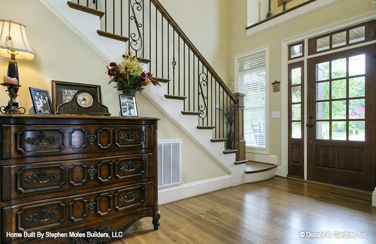 Foyer with an ornate staircase and an antique console table topped with a clock, framed photos, and a table lamp.