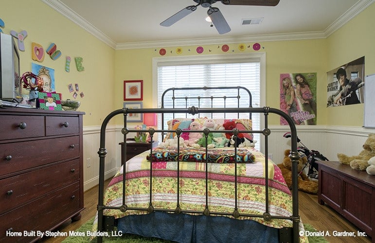 Another bedroom with a metal bed, wooden dresser and bench, and yellow walls adorned with various artworks and white wainscoting.