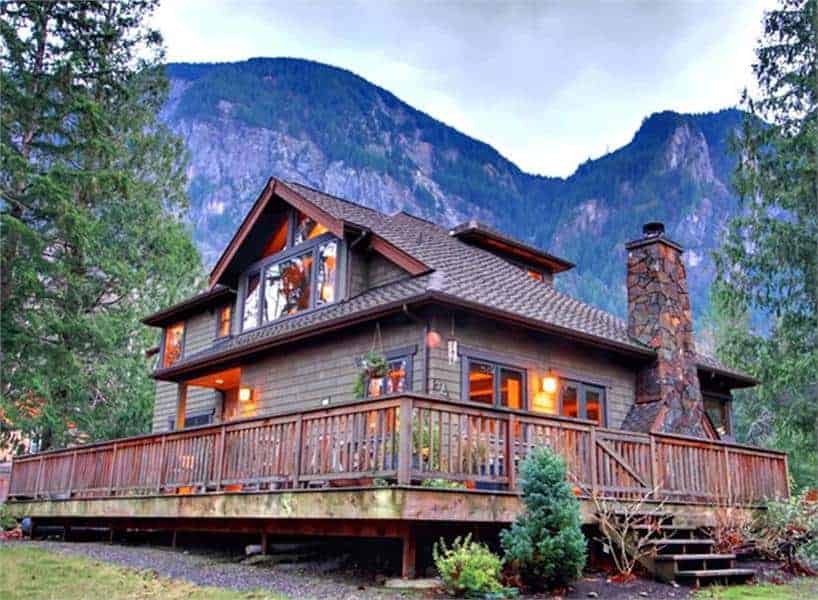 This is a rear view of the moutain-style home that has a wrap-around balcony porch with wooden railings and walkway. These are then complemented by the surrounding landscape of trees and shrubs.