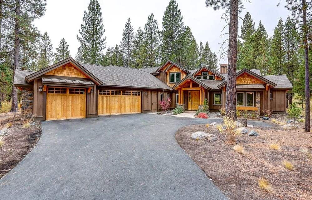 This is a view of the front of the mountain chalet-style home that has a wide asphalt driveway, wooden garage doors and landscaping of shrubs, trees and decorative rocks.