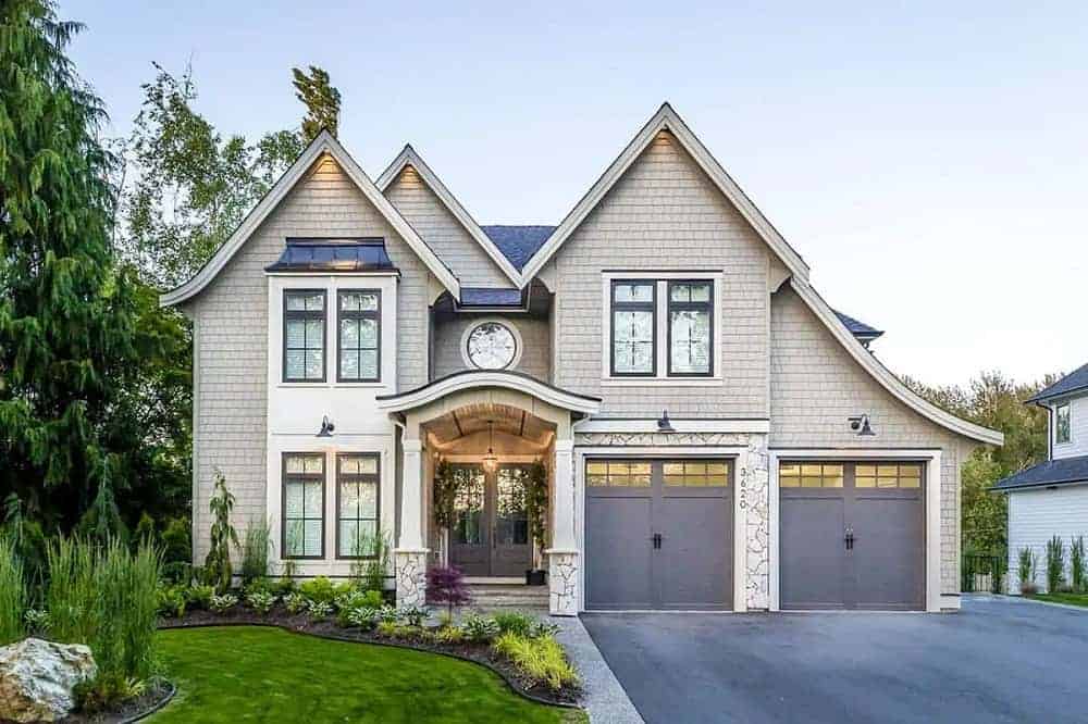 This is a front view of the house with multiple A-frame roofs, a large concrete driveway and a miniature front lawn garden with a small grass lawn, shrubs and small trees by the main entrance.