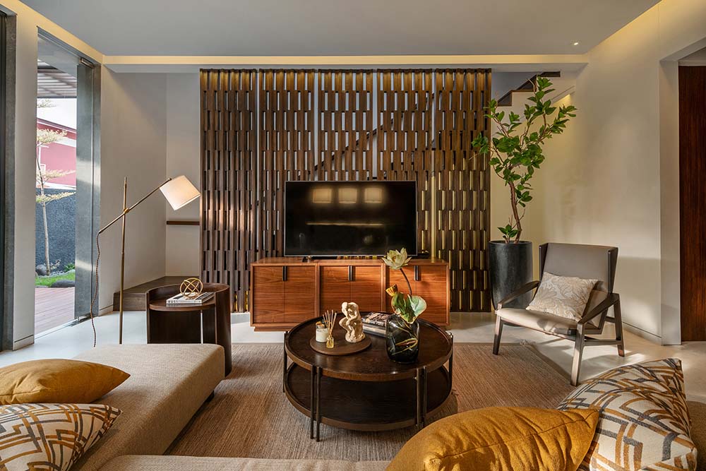 The living room of the house has a dark round coffee table in the middle of the brown area rug surrounded by armchairs and sofas across from the TV on an entertainment cabinet.