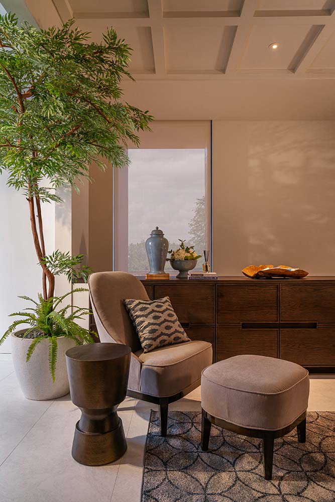 On the side of the cushioned chair and reading nook is the wooden waist-high cabinet adorned with a large potted plant and various decors on top.