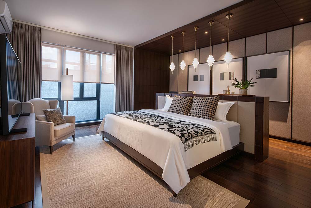 The primary bedroom has a large bedwith dark wooden accents that match the alcove with dark wooden elements complemented by the row of pendant lights and the wall-mounted artworks.