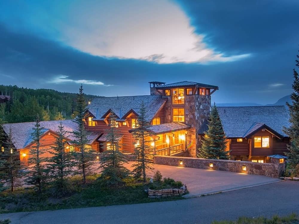 This is a view of the house exterior from the vantage of the large concrete driveway showcasing the warm glow of the house’s windows and balconies. These are then complemented by the landscaping of tall pine trees, mosaic stone walls and outdoor lights.