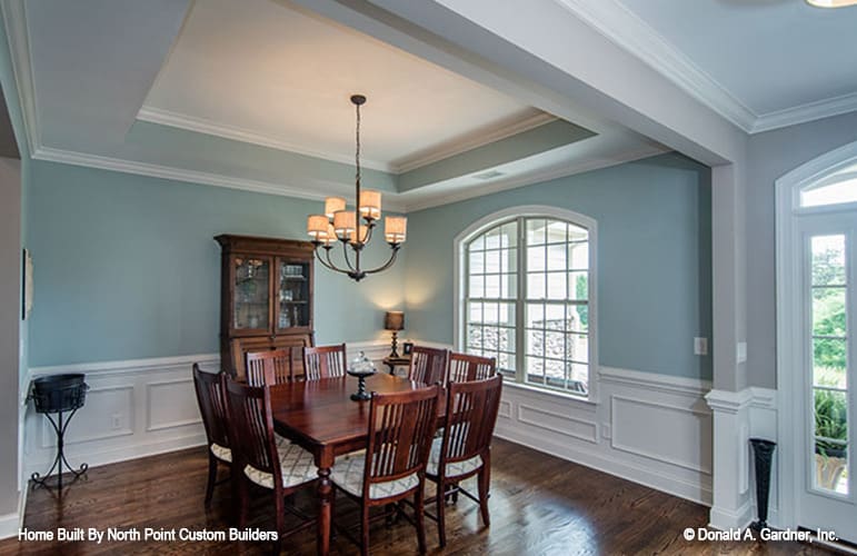 Dining area with dark wood furnishings, a tray ceiling, an arched window, and blue walls adorned with white wainscoting.