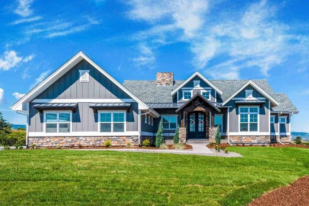 This is the front of the house adorned with multiple windows and gray shiplap exterior walls with stone mosaic base accents that go well with the shrubs planted on the sides of the concrete walkway and grass lawn.