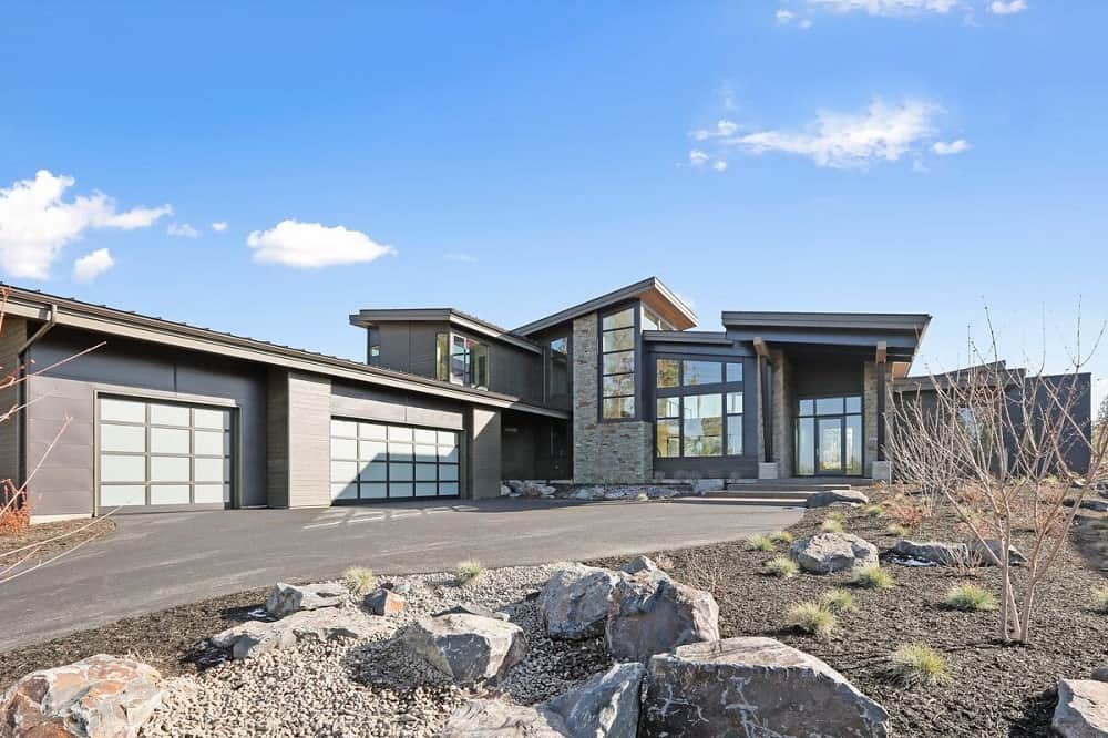 This is a view of the front of the house that has an abundance of glass walls and windows complemented by the concrete driveway, small dessert shrubs and large decorative rocks.