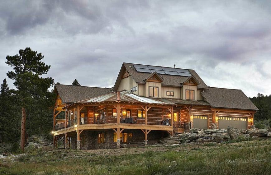 This is a view of the rear of the house that has a wrap-around porch supported by wooden beams below and complemented by the landscaping of grass, trees and large decorative rocks that bring a rustic quality to the house.
