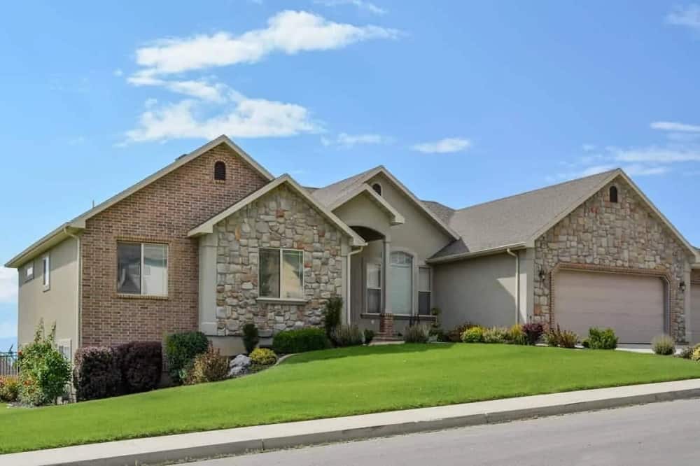 This is a view of the front of the house that has both red brick exterior walls and mosaic stone exterior walls complemented by the landscaping of grass lawn and colorful shrubs lining the sides.