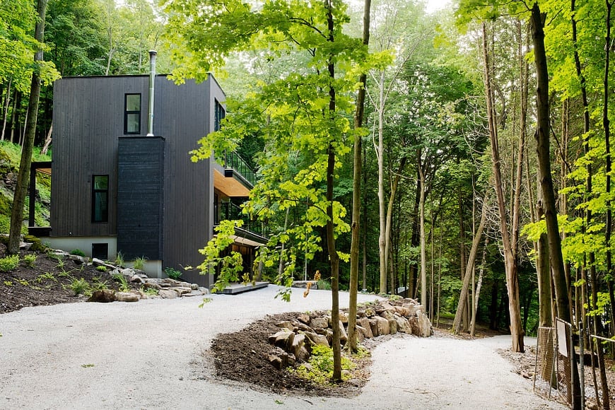 This is a far-off view of the chalet home surrounded by a lush landscape of graveled driveway, tall trees and stone structures.