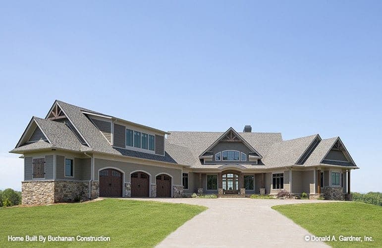 This is a front view of the house exterior from the vantage of the wide concrete driveway leading to a courtyard with planters and shrubs by the exterior walls. These are then complemented by the large grass lawn.