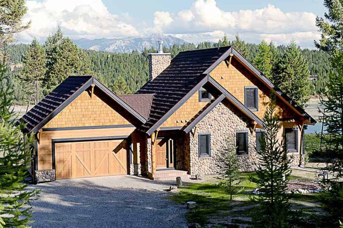 This is a front view of the house with agraveled driveway and walkway complemnted by the large grass lawn dotted with various pine trees that go well with the earthy tones of the house exterior.