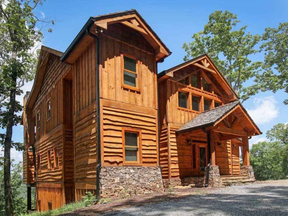 This is the front of the house that has consistent wooden textured walls adorned with windows and stone mosaic accents that go well with the graveled walkway and shrubs.