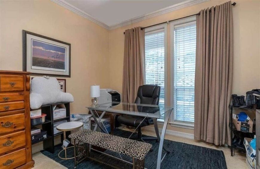 Study with large windows, wooden dresser, and a glass top desk flanked by a swivel leather chair and a cushioned bench.
