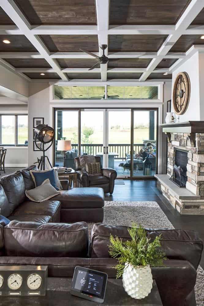 Great room with leather seats, a stone fireplace, and a coffered ceiling mounted with a fan.