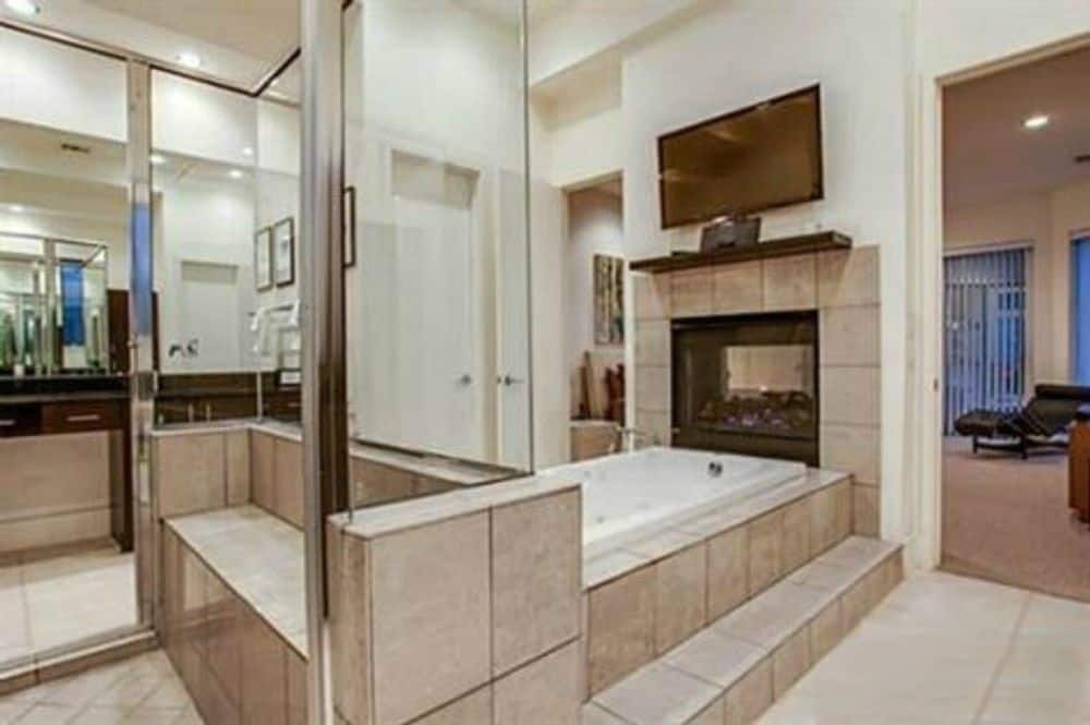 The primary bathroom features a deep soaking tub topped with a double-sided fireplace and a wall-mounted TV.