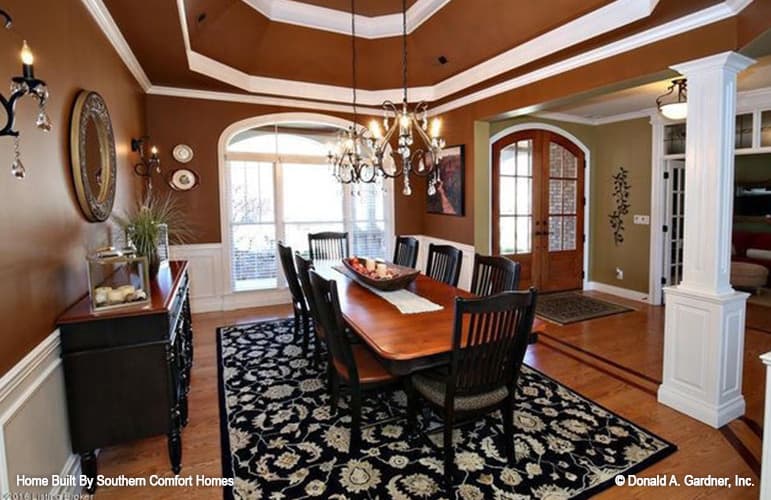 Dining room with an elegant step ceiling, dark wood furnishings, and brown walls graced with white wainscoting and various artworks.