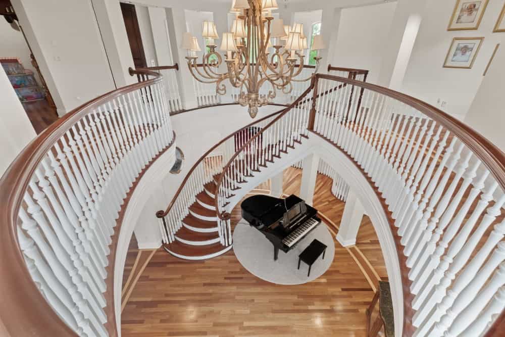 Top view of the soaring foyer from the upper landing showing the swirling staircases and a grand piano over a round rug. Image courtesy of Toptenrealestatedeals.com.