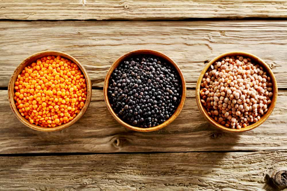 This is a close look at three bowls of various types of lentils on a wooden table.