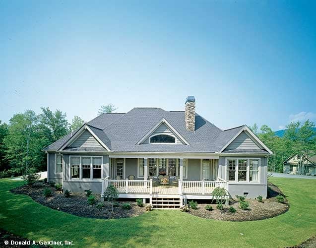 Rear exterior view with hipped and gable rooflines, a stone chimney, and an expansive covered porch bordered by white railings.