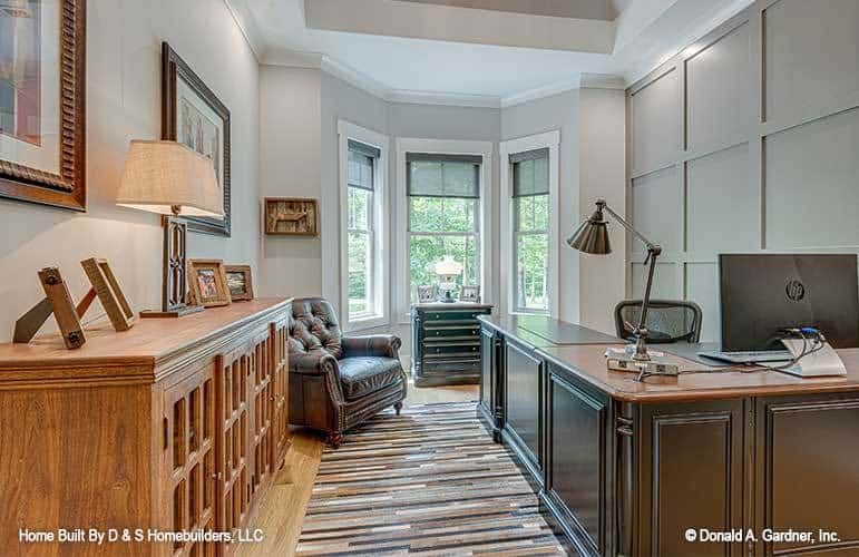 The study features a gray paneled accent wall, an L-shaped desk, a wooden cabinet, and a leather tufted chair sitting beside the bay window.