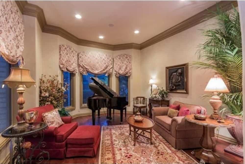 The living room is filled with a baby grand piano, comfy seats, a tall potted plant, and a bay window dressed in patterned roman shades.