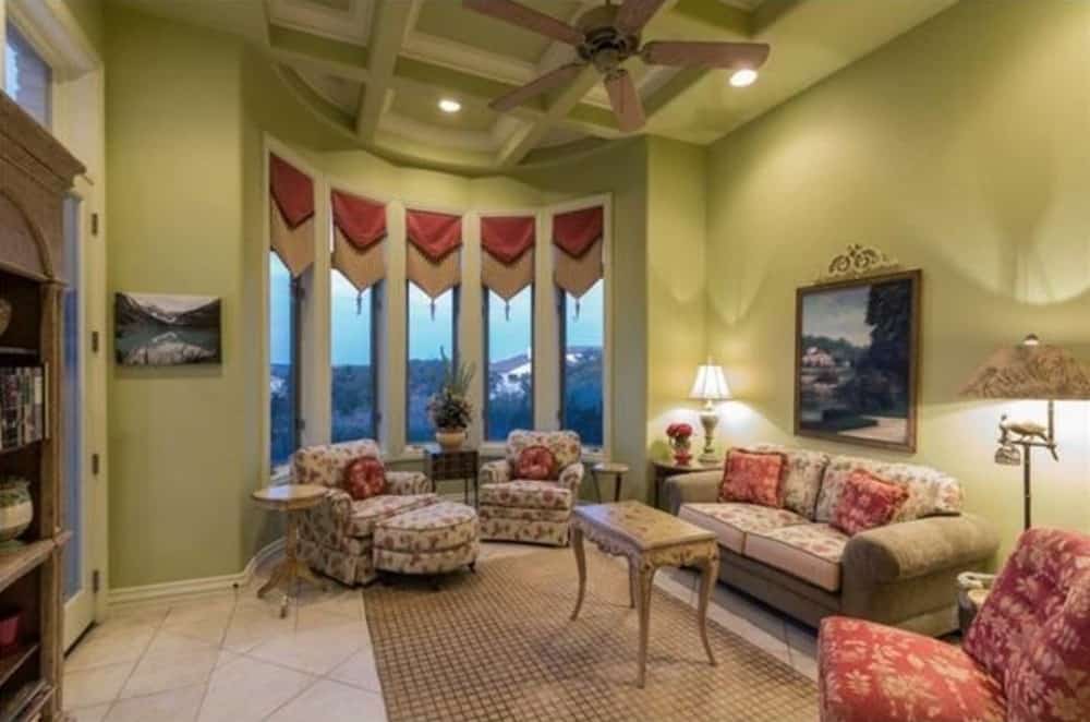 The library has patterned seats, wooden bookshelves, a bow window, and a coffered tray ceiling mounted with a fan and recessed lights.