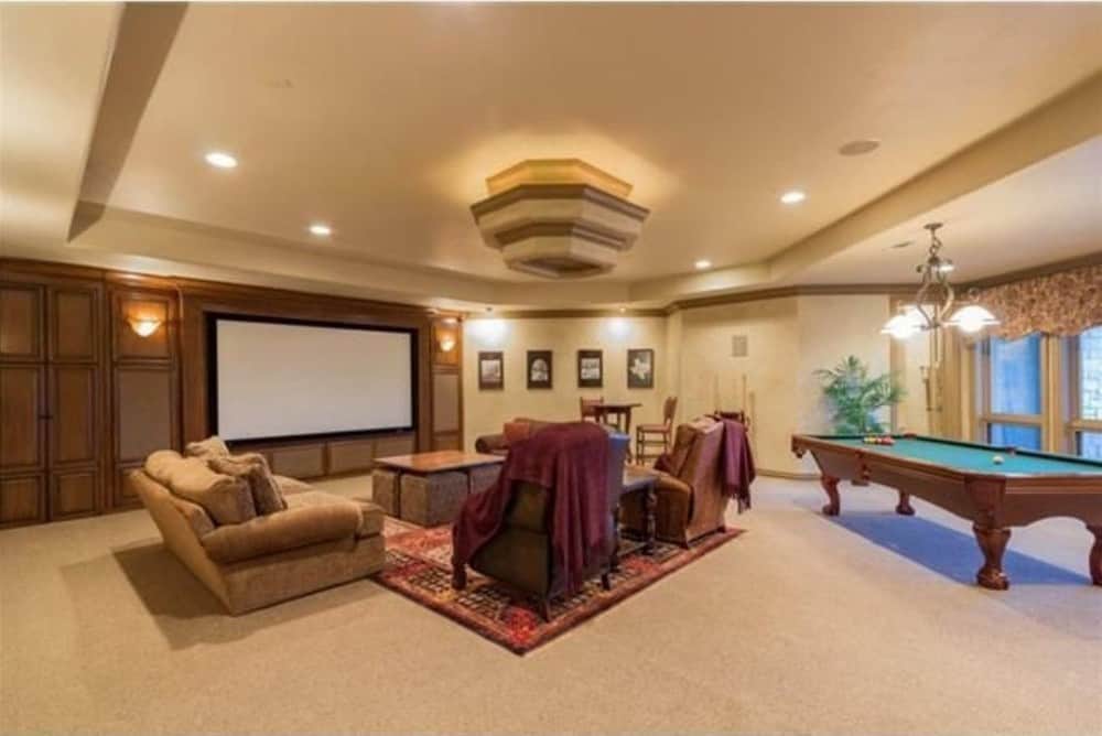 The recreation room is filled with fabric upholstered seats, a billiards table, and a large projector screen mounted against the wood-paneled accent wall.