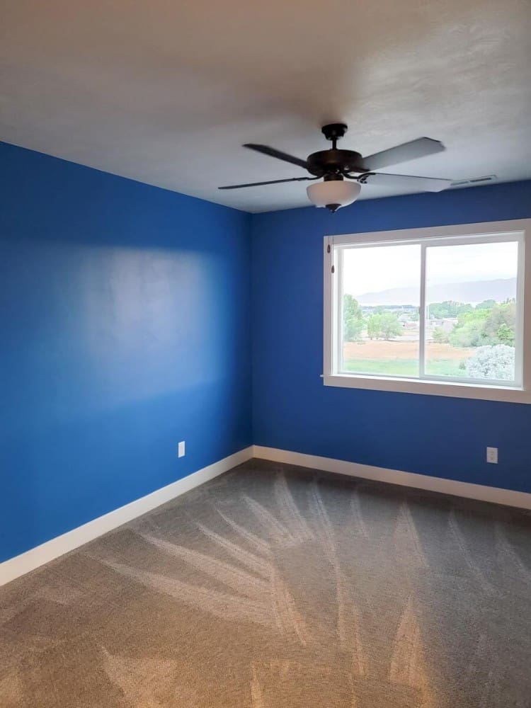 This bedroom has royal blue walls, an iron ceiling fan, and sliding glass windows overlooking the expansive surrounding.