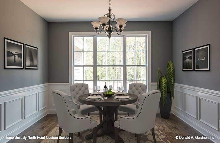 Formal dining room with an ornate iron chandelier, a round dining set, and gray walls adorned with white wainscoting and framed artworks.
