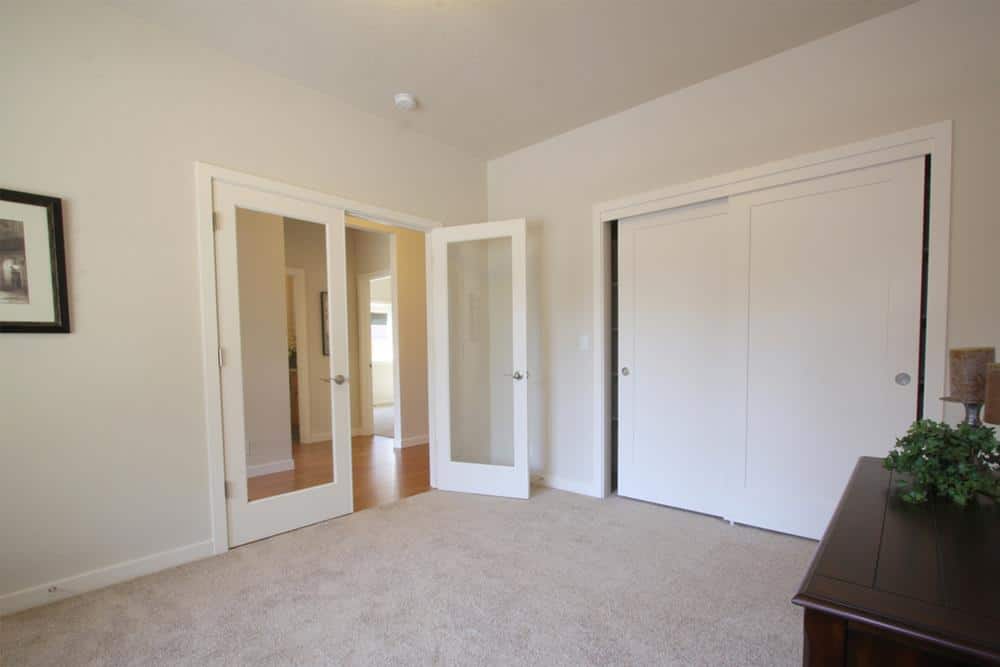 A french door opens to a den with carpet flooring, a dark wood desk, and a built-in wardrobe concealed behind the sliding white doors.